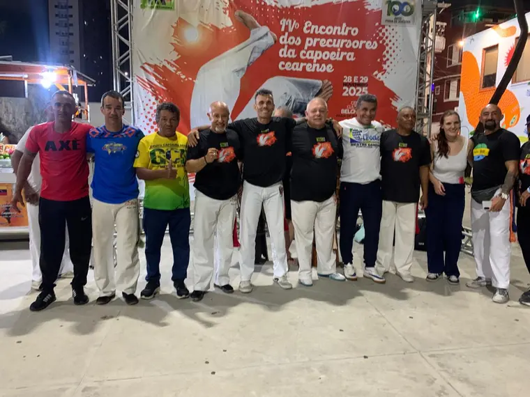 Grupo de capoeiristas posando ao lado do banner do IV Encontro dos Precursores