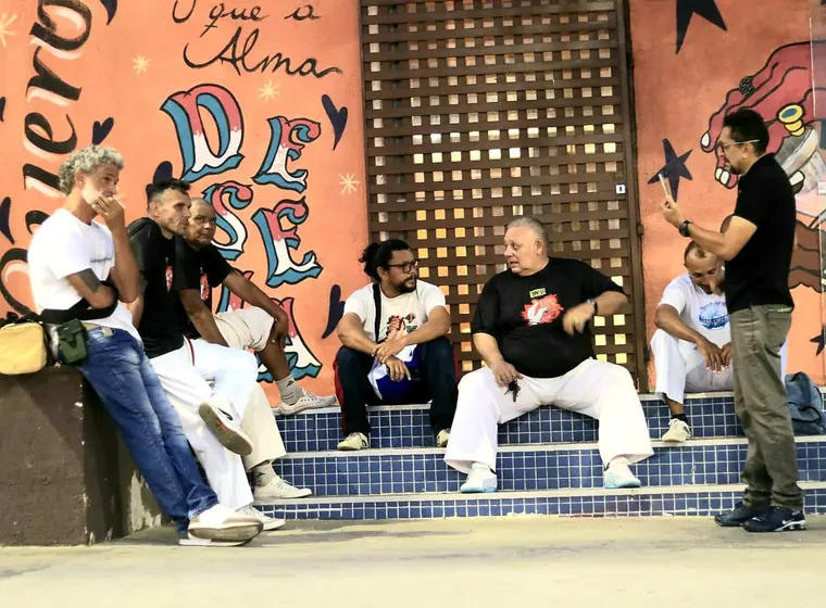 Três capoeiristas sentados em frente a um grafite colorido